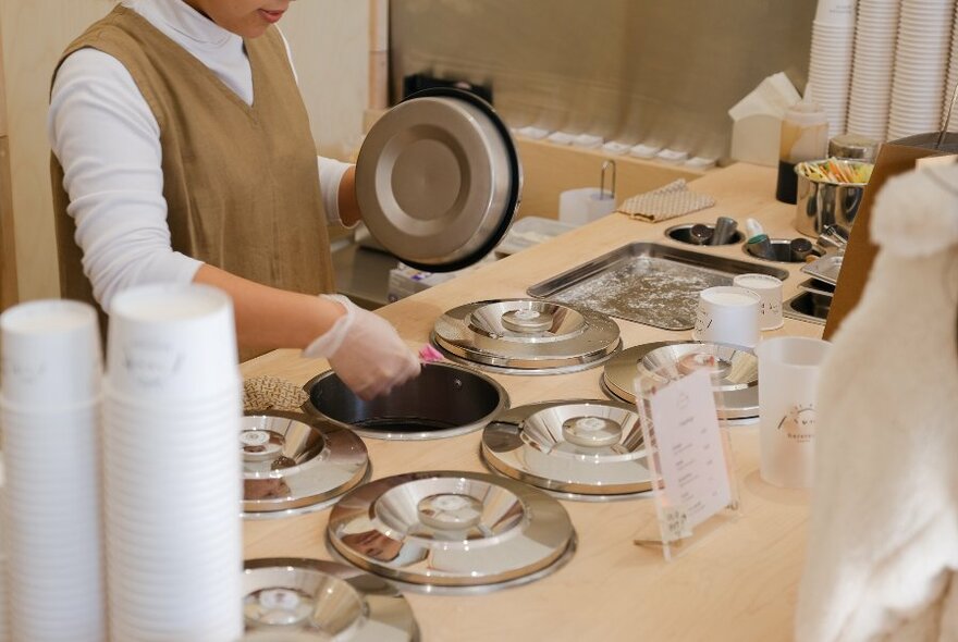 Person from chin down wearing white top and light brown shift, scooping gelato from bench with inset buckets with silver lids.