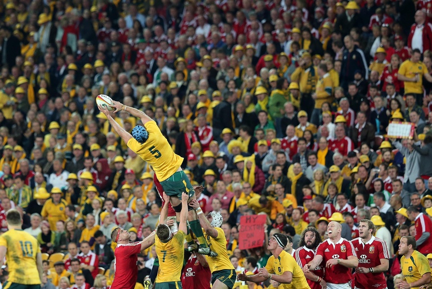 Rugby players from the Wallabies in yellow and green and from the Lions in red and white, playing rugby, with fans in the stadium cheering.