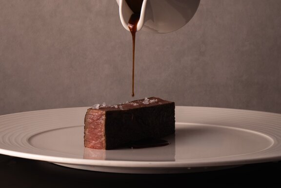 A perfectly rectangular piece of Wagyu steak in the centre of a white plate, gravy being poured onto it from a small, white gravy jug. 