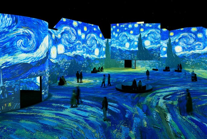 People walking around a very large space with Vincent van Gogh artwork digitally projected on all the wall and floor spaces.