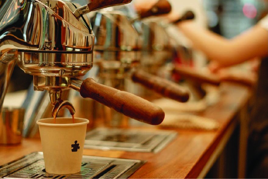 A professional espresso machine in a cafe setting dripping coffee into a paper takeaway cup.
