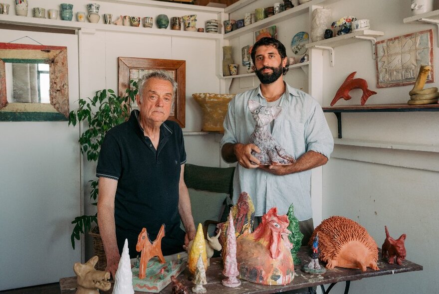 Two male sculptors standing next to a table of small animal sculptures in a room decorated with small pottery items on shelves.