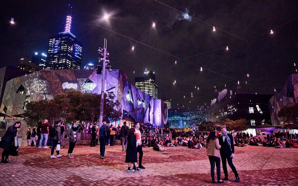 People milling around Fed Square at night.