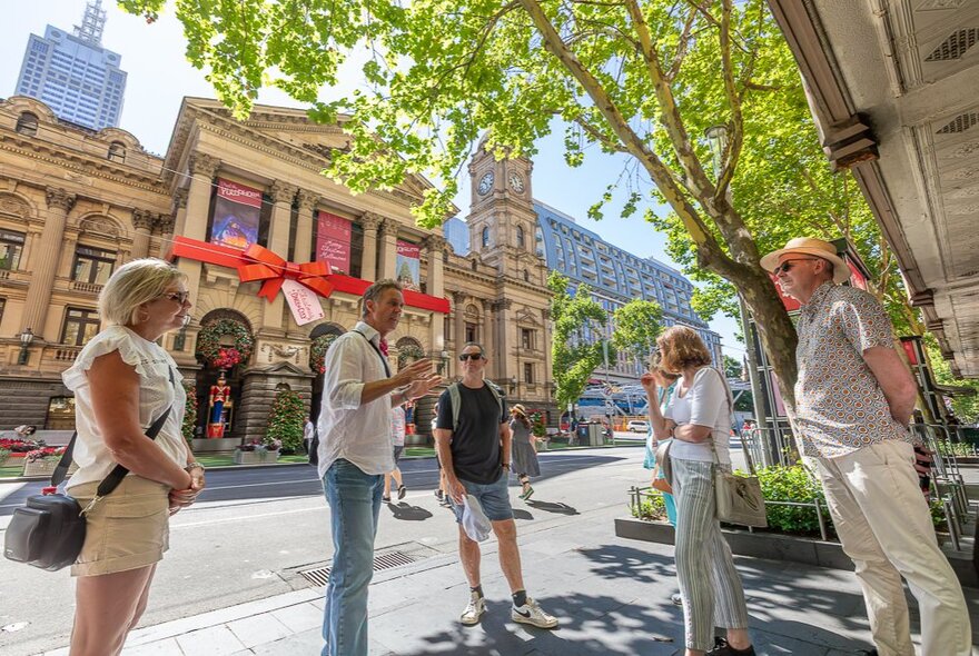 Small tour group standing under a tree in front of Melboune Town Hall, listening to a man speaking.