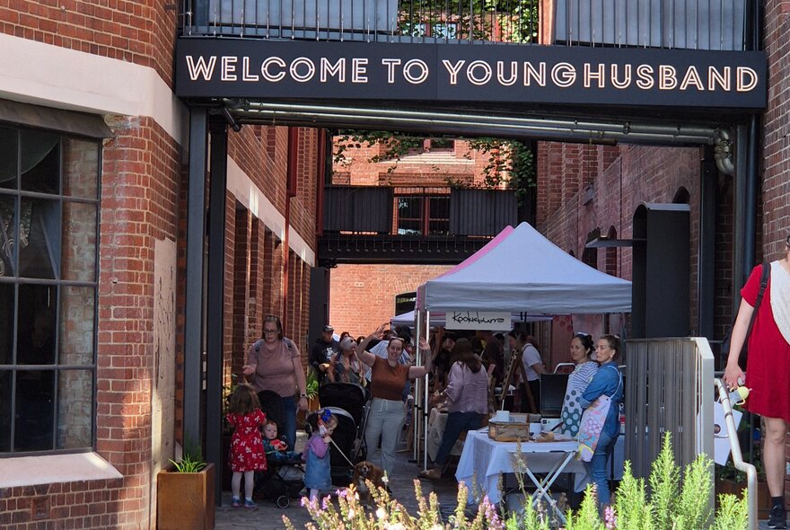 Market stall tents with signage next to a heritage brick building, with greenery and families.