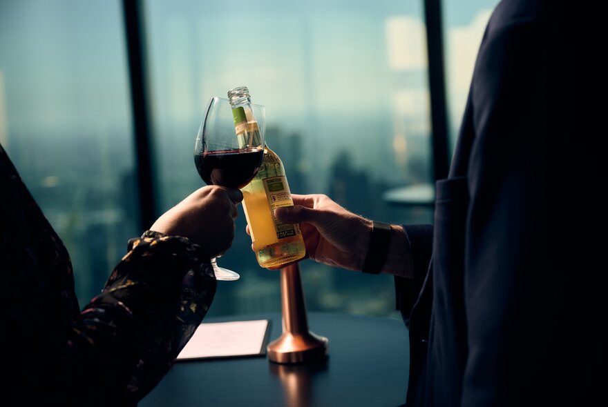 A person holding a glass of red clinks the glass against a person holding a stubbie of beer.