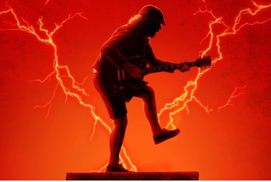 Angus Young silhouetted against a red lightning-struck sky, playing guitar in schoolboy shorts and cap.