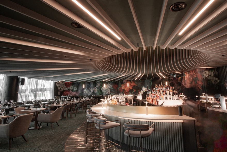 Interior of Amphora restaurant with rows of curved strip lighting on the ceiling leading to a central bar area, with dining tables and chairs arranged around the central bar.