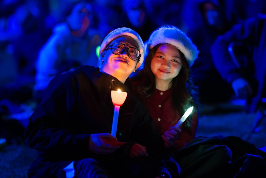 Two individuals holding torches under their chins that light up their faces against a dark crowd of people, during the festive community concert of Carols by Candlelight.