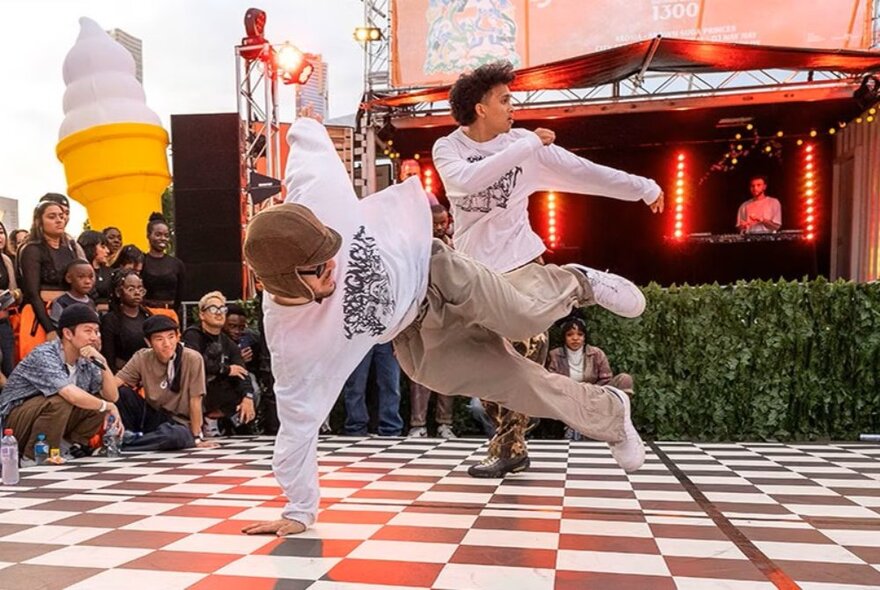 Two street dancers, dancing on a red and white check floor area outdoors ar Fed Square with people watching, and a DJ in the background playing music.