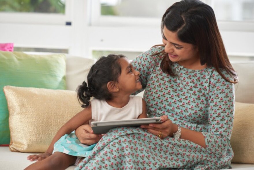 A woman with her arm around a little girl on a sofa reading her a book.