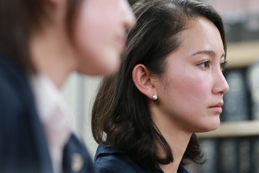 Film still; a profile of a young woman, her hair pushed back behind her ear, a pearl stud earring, with an out of focus head in the foreground in front of her.