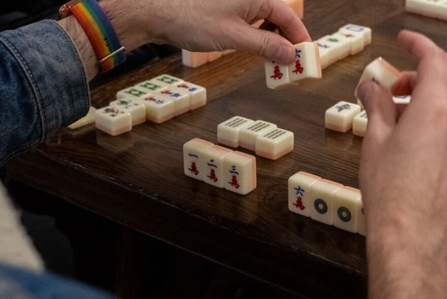 Hands arranging mahjong tiles on a table during a game.