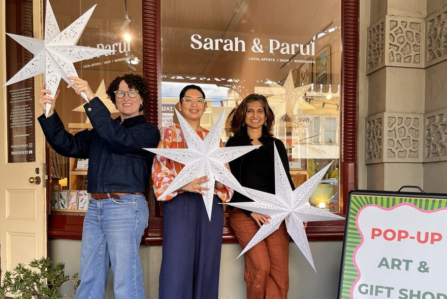 Three women posing in front of the store Sarah and Parul on Errol Street, holding up silver stars.