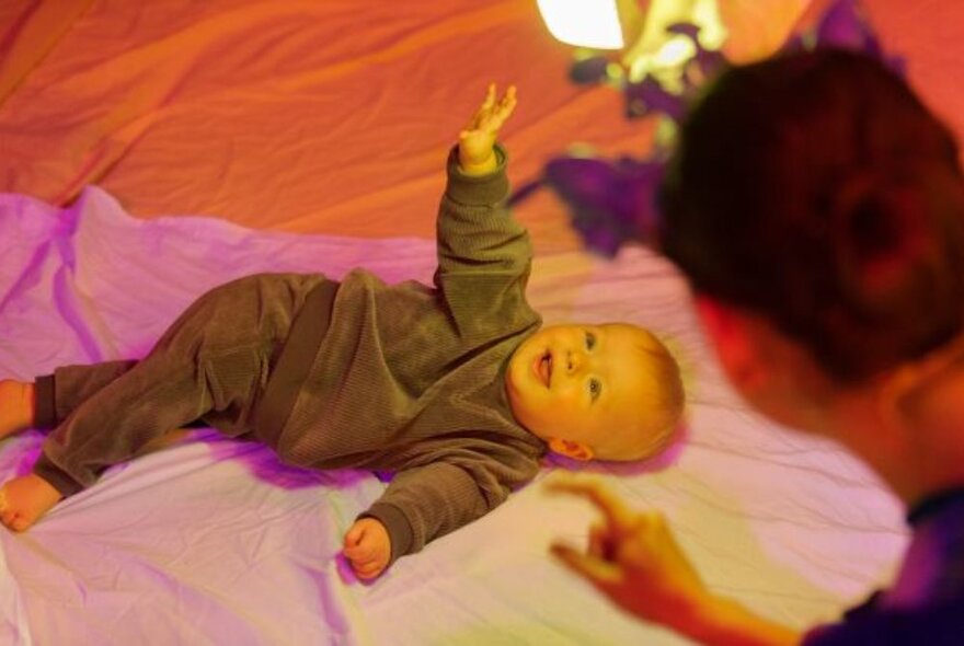 A smiling baby wearing a brown tracksuit, lying on multicoloured silk with an adult seen from behind in the foreground. 