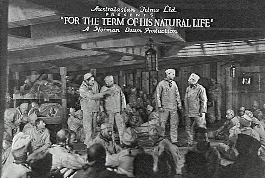 A black and white still from the Australian slient film, For the Term of his Natural LIfe, with four convicts standing amongst a large group of other convicts, all seated.