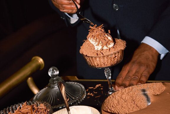 A waiter's hands sprinkling a garnish of shaved chocolate on a chocolate mousse served in a tall glass. 