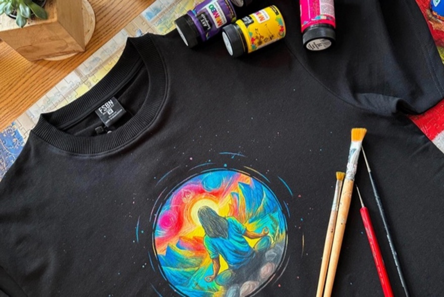 A hand-painted custom black t-shirt featuring a colourful circular artwork of a meditating figure with small pots of paint and brushes resting on the t-shirt.