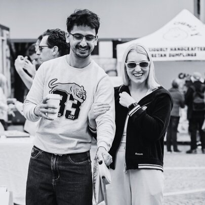 A black and white image of a couple smiling at an outdoor market, wearing sunglasses. 