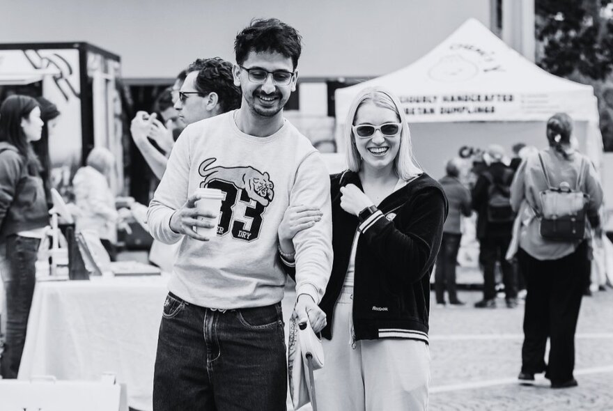 A black and white image of a couple smiling at an outdoor market, wearing sunglasses. 