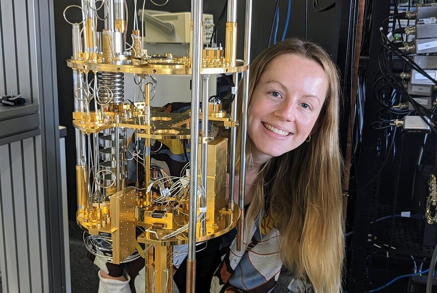 A smiling person with long blonde hair who is peeping her head out from behind a large brass scientific instrument.