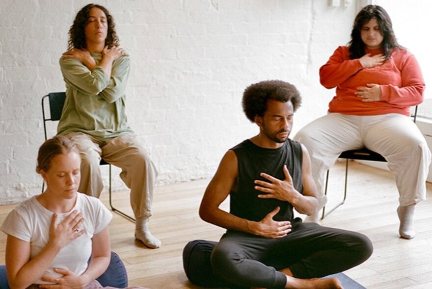 Four people in a wellbeing session, seated with hands on their chests and shoulders.