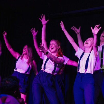 Ensemble cast of performers on stage, all wearing white t-shirts, blue trousers and braces, with their arms raised in the air, improvising in front of an audience. 