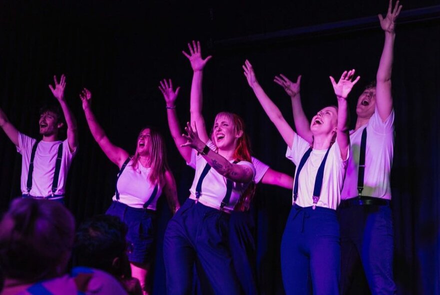 Ensemble cast of performers on stage, all wearing white t-shirts, blue trousers and braces, with their arms raised in the air, improvising in front of an audience. 