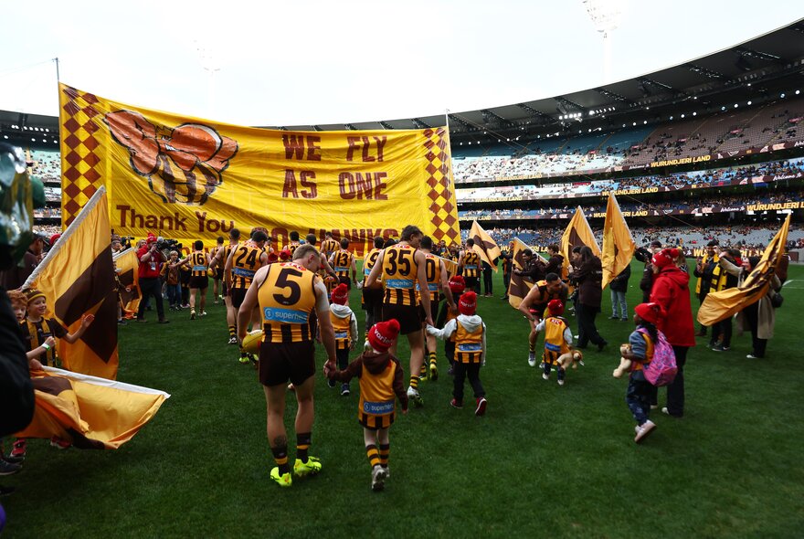 Hawthorn AFL players and their cheering squad running through the Hawthorn banner on the football field, with large crowds in the stadium in the background.
