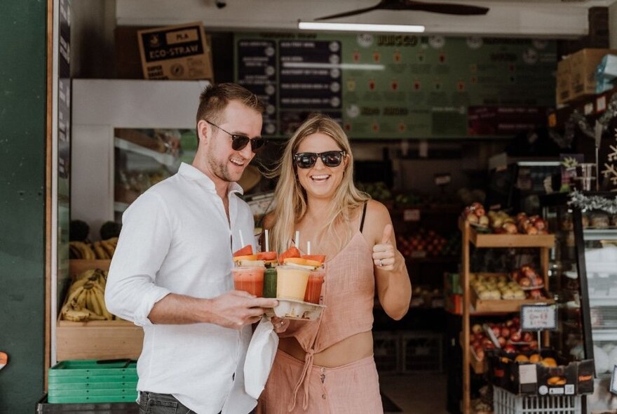 A couple walking out of a greengrocer store, smiling and both wearing sunglasses and holding takeaway drink containers in their hands.