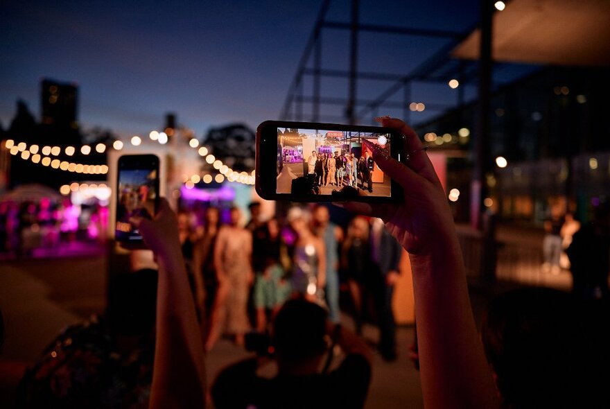 Night view of people gathered and taking photos using smartphones at Melbourne Museum Plaza during the Melbourne Fashion Festival.