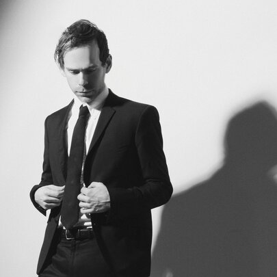 Composer Bryce Dessner dressed in a suit and looking downwards, his shadow visible on the wall. 