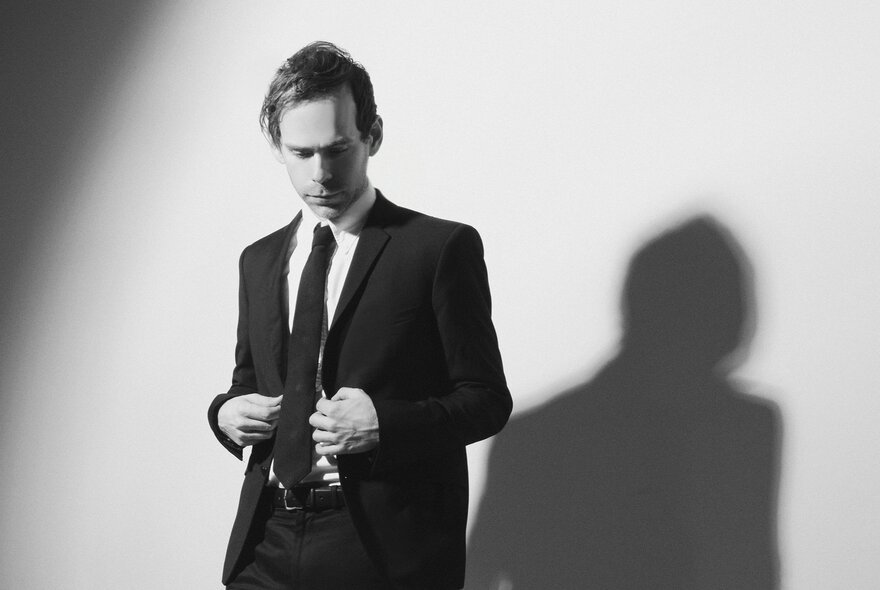 Composer Bryce Dessner dressed in a suit and looking downwards, his shadow visible on the wall. 
