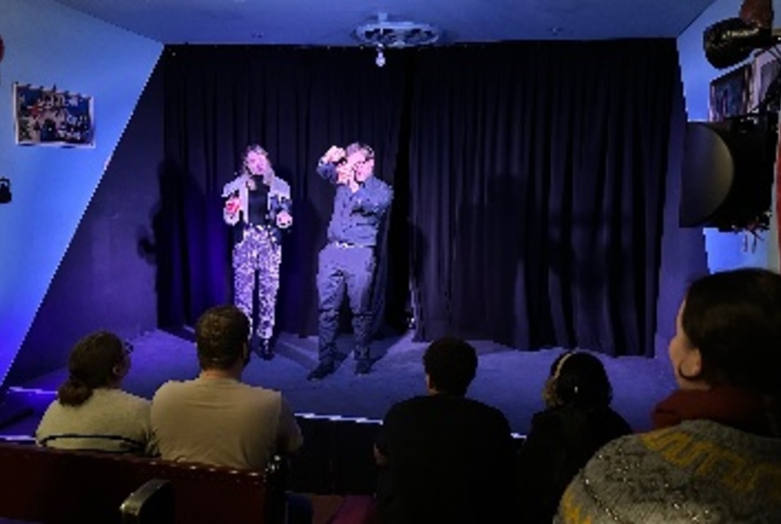 Two people on a stage in front of a black curtain, audience members seated in front of them.