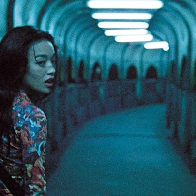 Movie still of a woman walking through an underground tunnel with arches and overhead lighting.