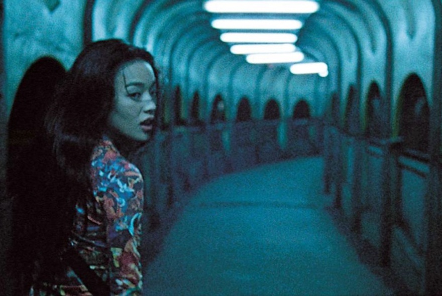Movie still of a woman walking through an underground tunnel with arches and overhead lighting.