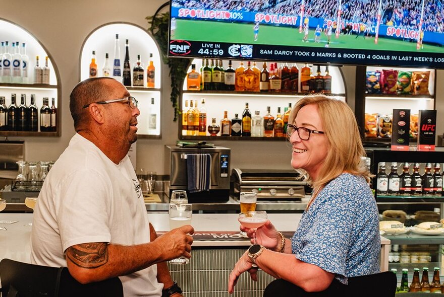 A bar setting with two people on stools and a vast array of drinks on shelves in the background. 