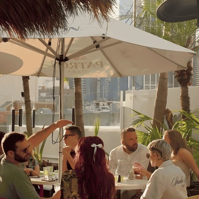 Patrons enjoying drinks under a cafe umbrella at a rooftop bar in the late summer evening.