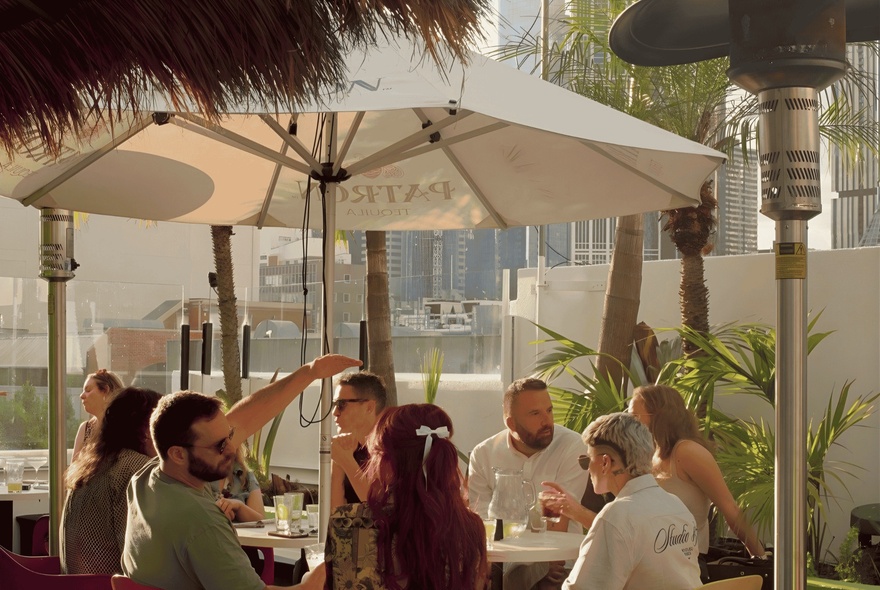 Patrons enjoying drinks under a cafe umbrella at a rooftop bar in the late summer evening.