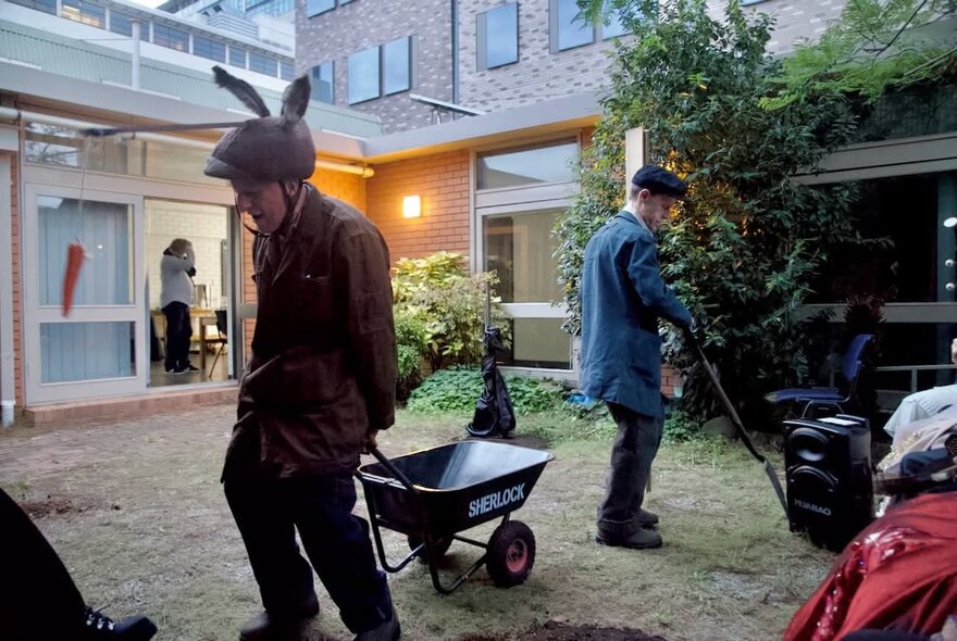 A scene in a courtyard garden with a man wearing a helmet with bunny ears and another gardening. 