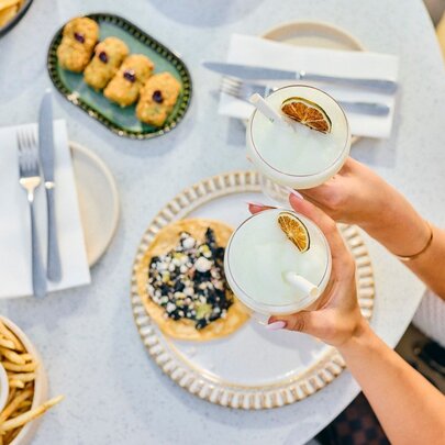 Two people holding margaritas, seen from above, with plates of Mexican food on a white table below.