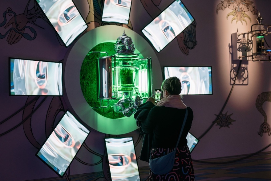 A person looking at artwork in a gallery space, the piece has a green glass centre and eight identical pictures arranged around it like petals on a flower. 