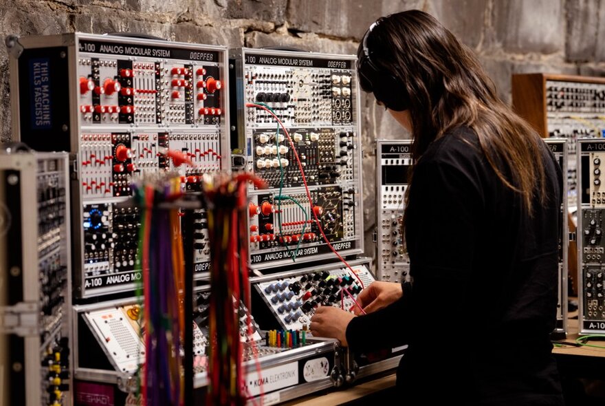A person wearing headphones, tweaking the knobs and controls on a large bank of electronic recording equipment.