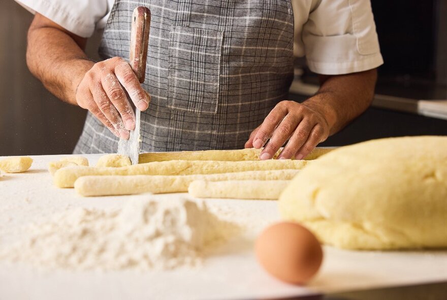 A torso shot of someone wearing a checked apron making gnocchi with a large mound of dough, an egg and some flour nearby. 