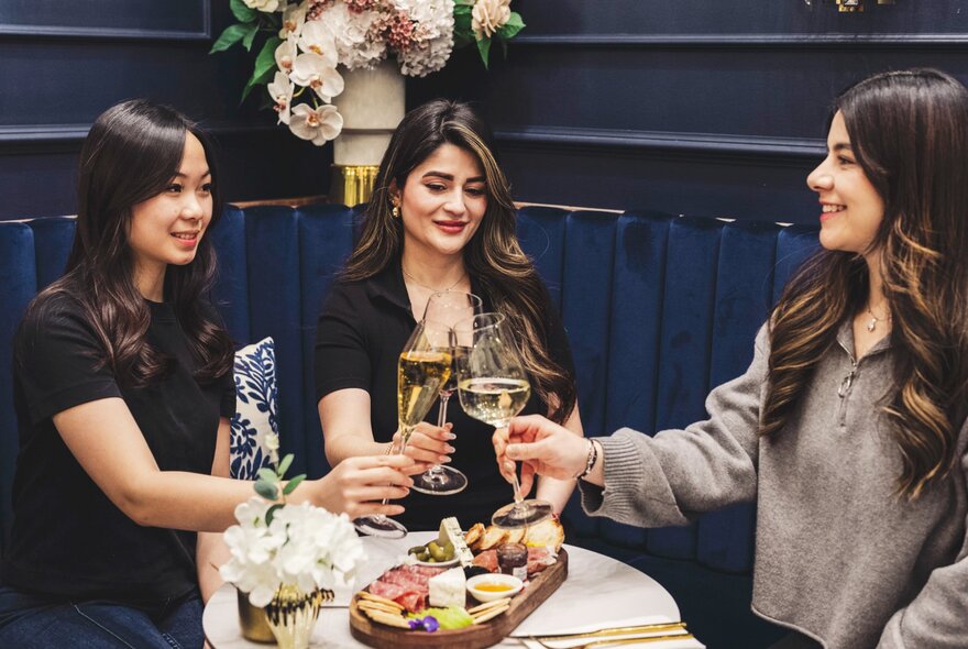 Three friends toasting with champagne and cocktails over a charcuterie board at a restaurant booth.