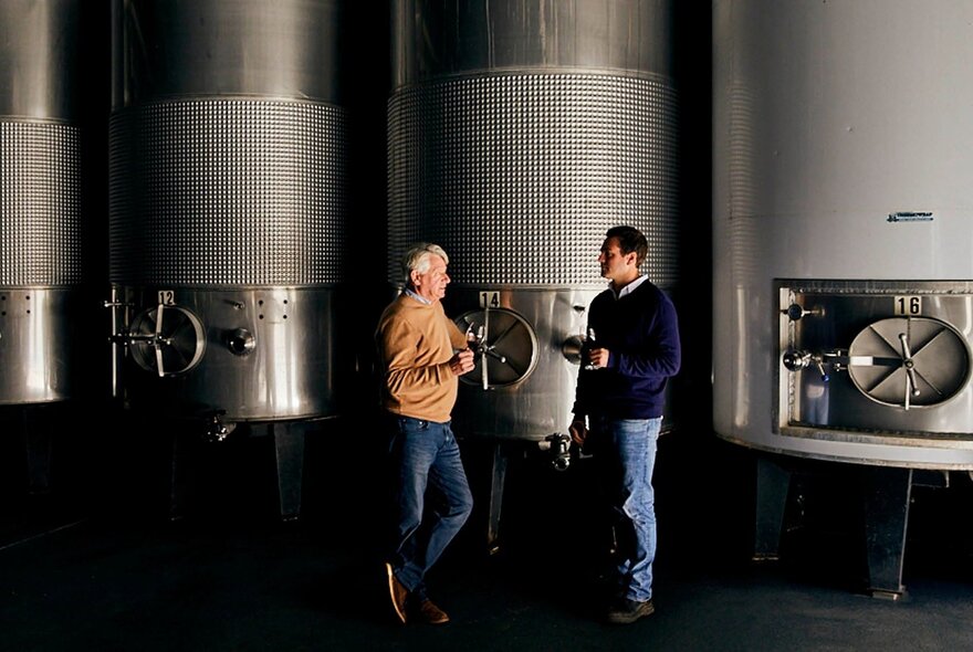 Two men standing with glasses of wine next to huge stainless-steel storage vats.