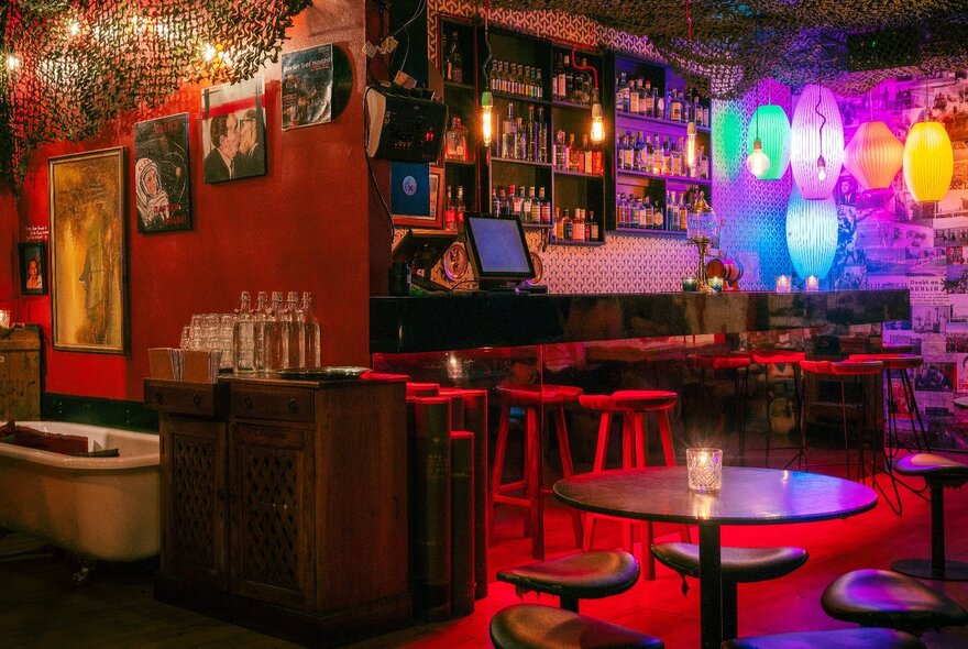 Interior of a bar decorated with colourful lamps, small paintings hanging on the red walls, with small tables and chairs arranged in the space.