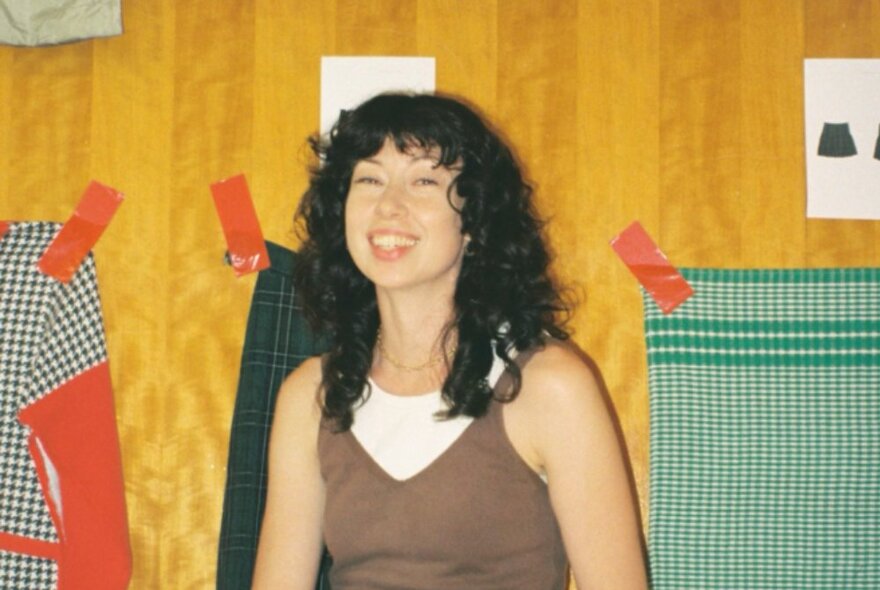 A smiling woman seated and visible from the waist upwards, behind them are checked tea towels taped to a wall.