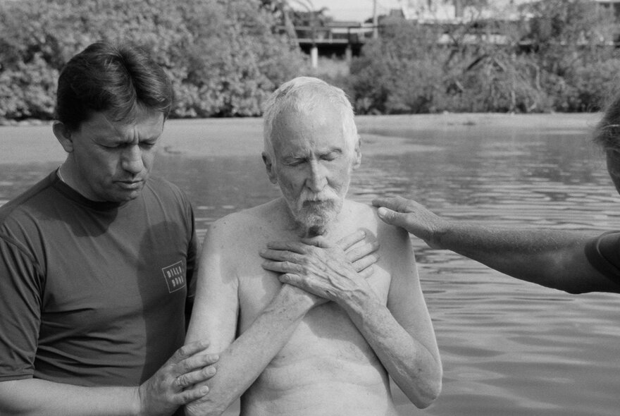 An elderly man being guided by another man in the sea, his hands crossed on his chest.