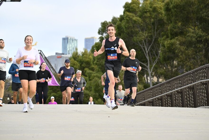 A group of people running over a bridge during a fun run, with trees and city buildings in the background.